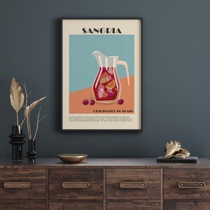 May include: A framed art print featuring a Sangria illustration. The artwork depicts a pitcher filled with red wine, fruit, and ice. Text at the top reads "SANGRIA," with "ORIGINATES IN SPAIN" below. The print is displayed on a wooden cabinet.