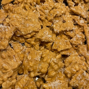 May include: Close-up of peanut brittle with a golden-brown color and a glossy finish. The brittle is broken into irregular pieces and sprinkled with white coconut flakes. The image shows a homemade dessert.