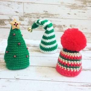 May include: Three crocheted Christmas decorations: a green Christmas tree with a gold star on top, a white and green striped elf hat, and a red and white striped hat with a red pom-pom.