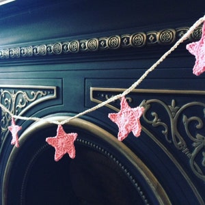 May include: A pink crocheted star garland hangs above a black fireplace. The garland is made of white yarn and features pink crocheted stars.