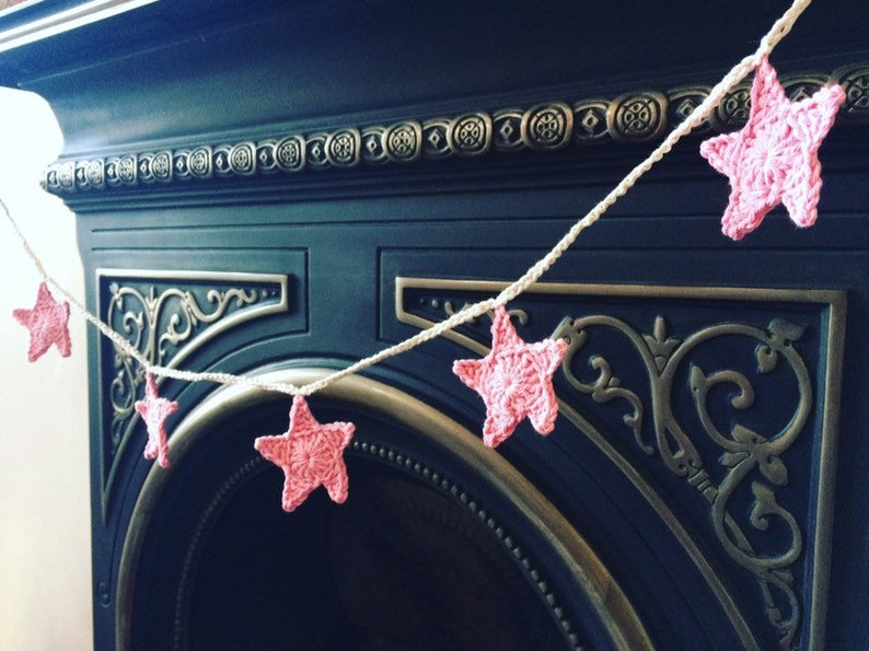 May include: A pink crocheted star garland hangs above a black fireplace. The garland is made of white yarn and features pink crocheted stars.