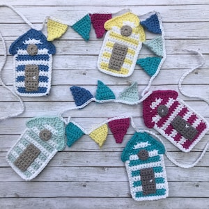 Op de afbeelding: Een kleurrijke gehaakte slinger met miniatuur strandhuisjes en vlaggen. De slinger is gemaakt van wit, blauw, geel, groen, roze en grijs garen.