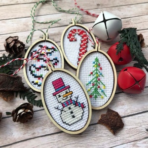 May include: Five wooden Christmas ornaments with different cross-stitch designs. The ornaments are round and have a white background. The designs include a snowman, a Christmas tree, a candy cane, and a string of lights.