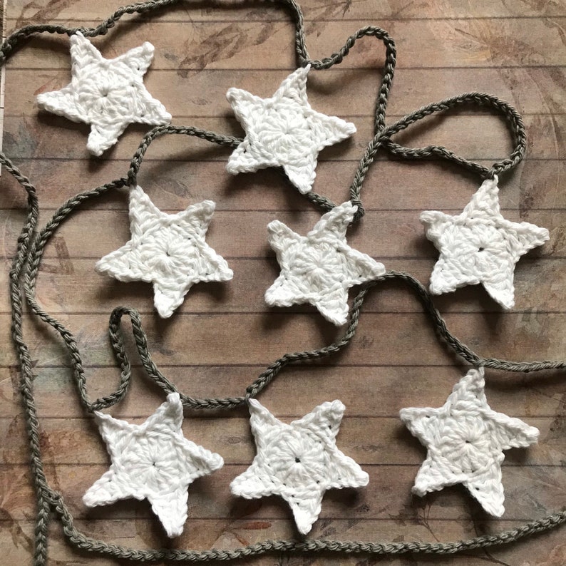 May include: A garland of white crocheted stars strung on a brown twine cord. The stars are evenly spaced and the garland is laid out on a woodgrain surface.