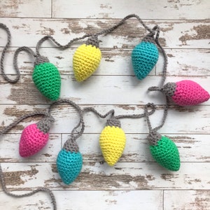 Könnte beinhalten: Eine Girlande aus gehäkelten Glühbirnen-Anhängern in Gelb, Rosa, Grün und Blau. Die Anhänger sind durch eine graue Schnur verbunden.