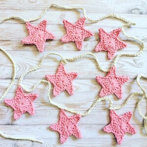 May include: A pink crocheted star garland with eight stars connected by a white string. The garland is hanging on a white wooden surface.
