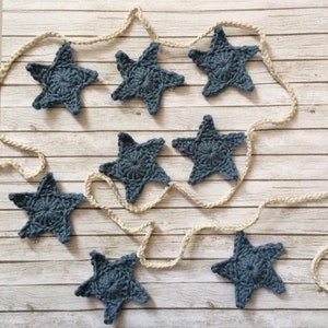May include: A garland of crocheted blue stars strung on a natural twine rope. The stars are a light blue color and have a simple, classic design.