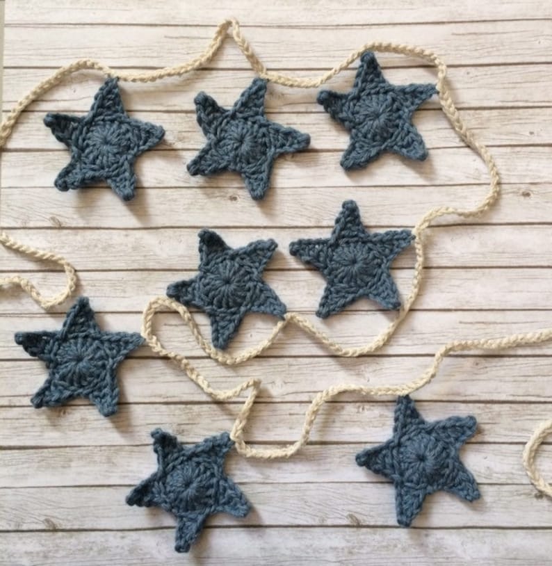 May include: A garland of crocheted blue stars strung on a natural twine rope. The stars are a light blue color and have a simple, classic design.