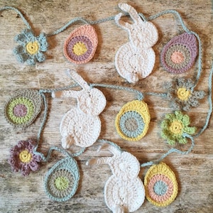 May include: A crocheted garland with white bunnies, pastel colored eggs, and flowers. The garland is made of a light blue yarn and is perfect for decorating for Easter.