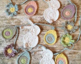 Op de afbeelding: Een gehaakte slinger met witte konijnen, pastelkleurige eieren en bloemen. De slinger is gemaakt van lichtblauw garen en is perfect voor Pasen decoratie.