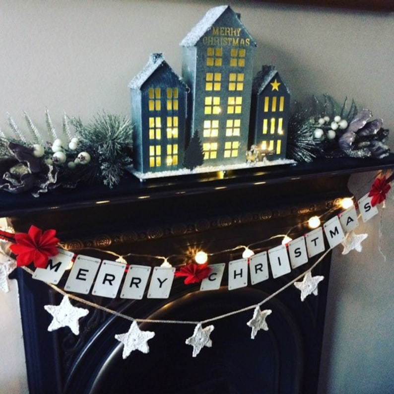 May include: A white and black banner with the words "Merry Christmas" spelled out on it. The banner is decorated with white crocheted stars and red felt flowers. A small, gray, wooden house with lit windows sits on a black mantelpiece behind the banner.
