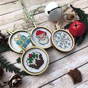 Op de afbeelding: Vijf kleine ronde houten ornamenten met verschillende kerstthema cross-stitch ontwerpen. De ornamenten zijn rond en hebben een witte achtergrond met kleurrijke borduurwerk. De ontwerpen omvatten een sneeuwvlok, de kerstman, een kerstpudding, een geschenkdoos en een kerstboom.