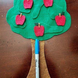 May include: A felt green tree with a brown felt trunk and five red felt apples with green felt stems and leaves. A blue marker is leaning against the trunk.