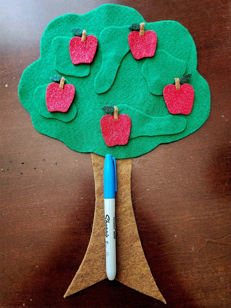 May include: A felt green tree with a brown felt trunk and five red felt apples with green felt stems and leaves. A blue marker is leaning against the trunk.