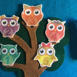 May include: Five felt owls with numbers 1 through 5 on a brown tree trunk with green leaves. The owls are different colors: purple, orange, red, green, and yellow.