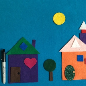 May include: Felt craft depicting two houses with a green tree and a yellow sun. The house on the left has a purple body, green roof, a red heart, and a brown door. The house on the right has an orange body, pink roof, a blue square, and a green door.