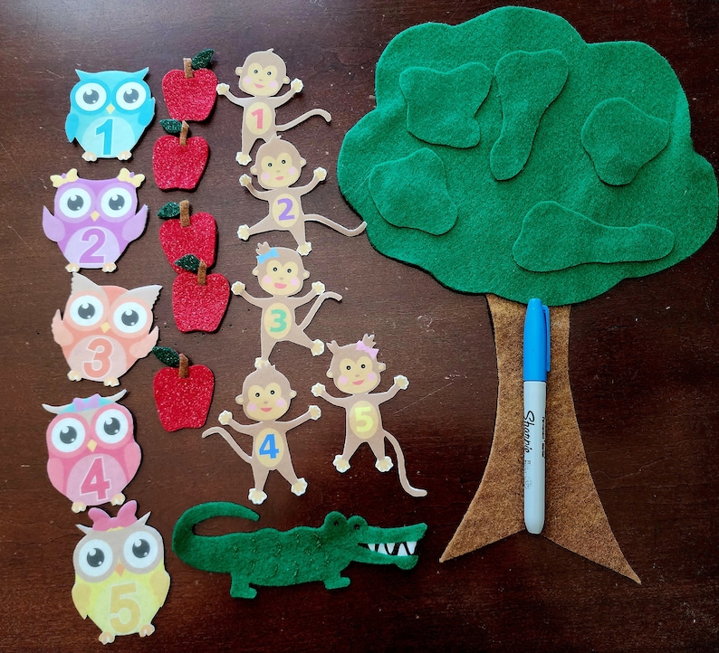 May include: A felt craft set for teaching counting with a green tree, five monkeys, five owls, and five red apples. The numbers 1 through 5 are on the owls and monkeys.