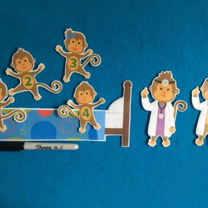K&ouml;nnte beinhalten: F&uuml;nf braune Filz-Affenfiguren mit den Zahlen 1 bis 4 auf ihren Hemden. Zwei Affen tragen Arztkost&uuml;me mit einem Stethoskop und einem Stirnband. Die Affen befinden sich auf einem blauen und wei&szlig;en Filzbett mit einem braunen Kopfteil. Ein schwarzer Sharpie-Marker befindet sich im Vordergrund.