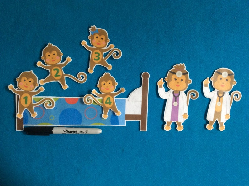 K&ouml;nnte beinhalten: F&uuml;nf braune Filz-Affenfiguren mit den Zahlen 1 bis 4 auf ihren Hemden. Zwei Affen tragen Arztkost&uuml;me mit einem Stethoskop und einem Stirnband. Die Affen befinden sich auf einem blauen und wei&szlig;en Filzbett mit einem braunen Kopfteil. Ein schwarzer Sharpie-Marker befindet sich im Vordergrund.