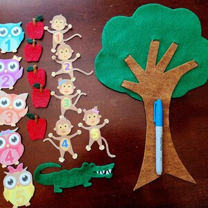 May include: A felt craft set for teaching children about numbers. The set includes a green felt tree with a brown trunk, felt owls with numbers 1 through 5, felt monkeys with numbers 1 through 5, felt apples, and a blue marker.