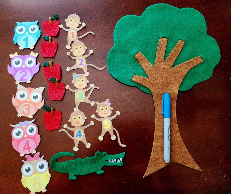 May include: A felt craft set for teaching children about numbers. The set includes a green felt tree with a brown trunk, felt owls with numbers 1 through 5, felt monkeys with numbers 1 through 5, felt apples, and a blue marker.