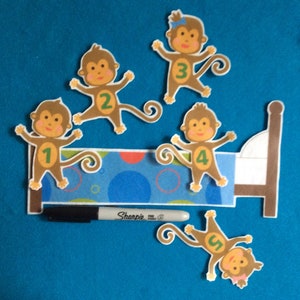 K&ouml;nnte beinhalten: F&uuml;nf braune Filz-Affenpuppen mit den Zahlen 1 bis 5 auf ihren B&auml;uchen. Die Affen sind um ein braunes Filzbett mit einem blau-wei&szlig; gemusterten Kopfteil angeordnet. Ein schwarzer Sharpie-Marker befindet sich im Vordergrund.