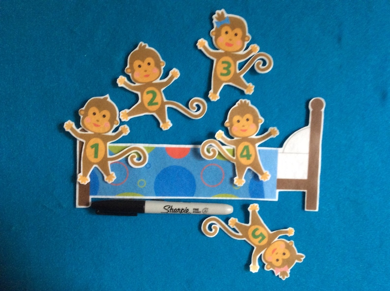 K&ouml;nnte beinhalten: F&uuml;nf braune Filz-Affenpuppen mit den Zahlen 1 bis 5 auf ihren B&auml;uchen. Die Affen sind um ein braunes Filzbett mit einem blau-wei&szlig; gemusterten Kopfteil angeordnet. Ein schwarzer Sharpie-Marker befindet sich im Vordergrund.