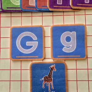 May include: Educational alphabet tiles with a blue background and orange border. The tiles feature the letters "G" and "g", and a giraffe illustration with the word "giraffe". The tiles are arranged on a grid.