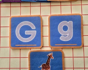 Puede incluir: Azulejos educativos del alfabeto con fondo azul y borde naranja. Los azulejos presentan las letras "G" y "g", y una ilustración de jirafa con la palabra "giraffe". Los azulejos están dispuestos sobre una cuadrícula.