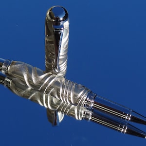 May include: A silver pen with a textured, swirling design. The pen has a chrome cap and a silver barrel. The pen is lying on a blue surface.