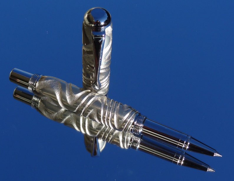 May include: A silver pen with a textured, swirling design. The pen has a chrome cap and a silver barrel. The pen is lying on a blue surface.