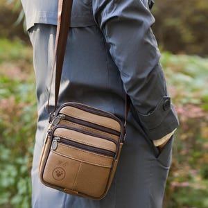 May include: A tan leather crossbody bag with three zippered compartments and a brown strap. The bag has a paw print emblem and the initials "M.C." on the front. The bag is worn over the shoulder.