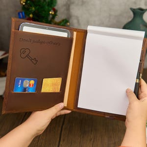 May include: Brown leather portfolio with a notepad, pen, and card slots. The portfolio has the text "Don't judge others" and a key graphic. A tablet is visible in the pocket. The portfolio is open, revealing a blank notepad.