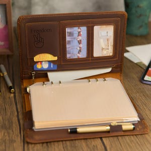 May include: Open brown leather binder with the word "Freedom" and a peace sign embossed on the cover. The binder has card slots, a zippered pocket, and a clear pocket for photos. A gold pen rests inside the binder.