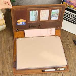 May include: A brown leather portfolio with multiple compartments, including card slots, photo holders, and a zippered pocket. The word "Brave" is embossed on the front. The portfolio is open, revealing a notepad and a pen.