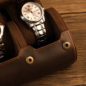 Peut inclure: Un porte-montres en cuir marron avec deux compartiments pour montres. Le porte-montres a deux boutons-pression en laiton et une montre argent&eacute;e avec un cadran blanc est visible dans l'un des compartiments.