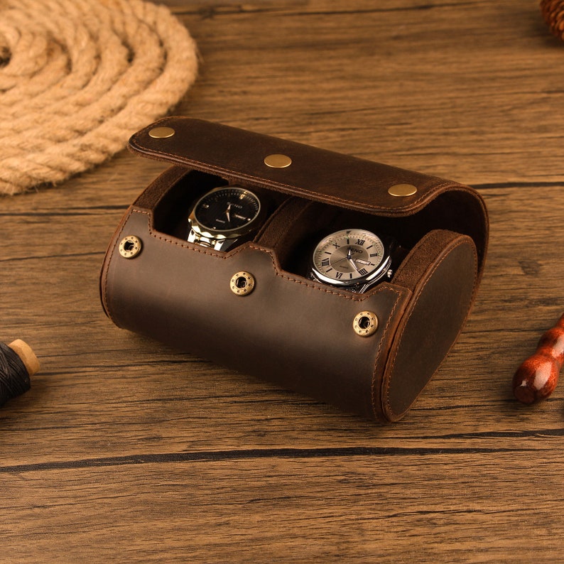 Peut inclure: Un porte-montres en cuir marron avec deux compartiments pour montres. Le porte-montres est ouvert et deux montres argent&eacute;es sont visibles &agrave; l'int&eacute;rieur.