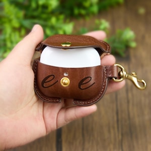 Puede incluir: Una funda de cuero marrón para AirPod con cierre de botón a presión y mosquetón dorado. La funda está abierta, revelando auriculares blancos. La funda tiene las letras "e e" en relieve.