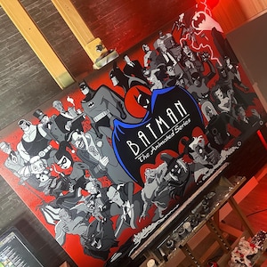 May include: A large, rectangular painting featuring characters from "Batman: The Animated Series." The artwork showcases Batman, the Joker, Harley Quinn, and other villains against a red backdrop. The title "Batman The Animated Series" is prominently displayed.
