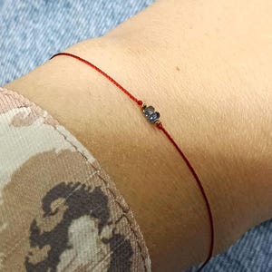 May include: A delicate bracelet featuring a thin red cord and two small, dark, faceted beads centered on the string. The minimalist design consists of a single strand of red thread that encircles the wrist, highlighting the tiny dark stones.