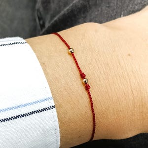 May include: A delicate red string bracelet featuring two small, polished gold-tone spherical beads spaced along the thin cord. The bracelet is tied with a simple knot, creating a minimalist look that rests against the skin on the wrist.