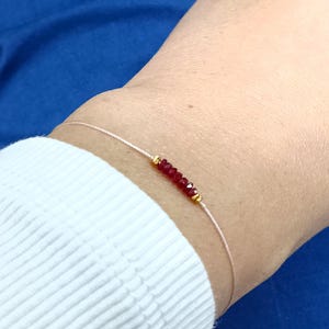 May include: Delicate bracelet featuring small, vibrant red beads strung on a thin, light-colored cord. The bracelet includes small gold-colored accents. The jewelry is shown on a wrist, against a blue background.