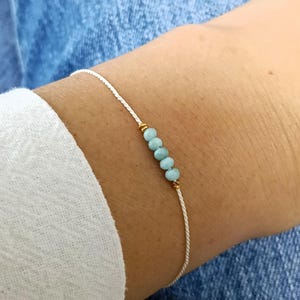 May include: A delicate bracelet featuring a thin white cord string. The center of the bracelet is adorned with four small, round, light blue beads arranged in a row, flanked by two tiny gold-toned metallic spacers on either side of the bead cluster.