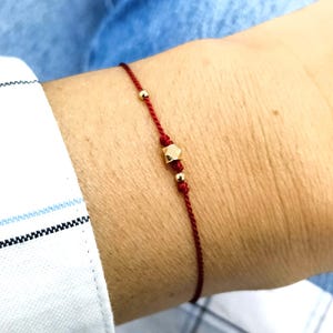 Puede incluir: Una pulsera de hilo rojo fina que presenta una pequeña cuenta cúbica dorada y facetada en el centro, flanqueada por dos diminutas cuentas redondas doradas a cada lado. El delicado cordón está atado con un nudo sencillo y elegante.