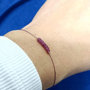Op de afbeelding: Een delicate armband met een reeks kleine, helderrode edelstenen. De edelstenen zijn gegroepeerd, waardoor een focuspunt ontstaat op het dunne, donkergekleurde koord. De armband wordt om een pols gedragen, met een witte mouw en blauwe stof op de achtergrond.
