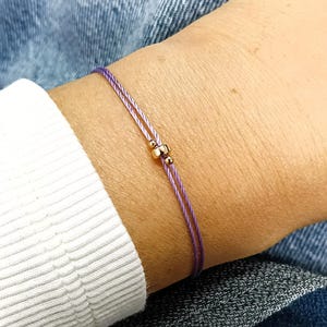 Może przedstawiać: Delikatna bransoletka składająca się z dwóch cienkich fioletowych sznurków. Bransoletka jest ozdobiona pojedynczym, małym, polerowanym metalowym koralikiem w złotym odcieniu, umieszczonym centralnie na dwuwarstwowym sznurku, co nadaje jej prosty wygląd.
