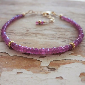 May include: A delicate bracelet featuring small, faceted pink gemstones strung together. Gold-coloured beads and a gold chain with an adjustable clasp complete the design. The bracelet is displayed on a light-coloured, textured wooden surface.