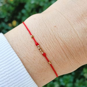 Puede incluir: Una delicada pulsera de hilo rojo que presenta una serie de pequeñas cuentas metálicas de tono dorado dispuestas en el centro, aseguradas por dos pequeños nudos a cada lado del grupo de cuentas, con cuentas doradas adicionales a lo largo.
