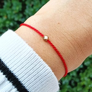 May include: A thin red cord bracelet featuring a single small, faceted gold-toned bead centred on the string. The bracelet is worn on a wrist, partially covered by a white ribbed sleeve with a black horizontal stripe near the cuff.