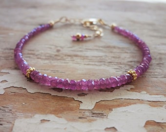 Op de afbeelding: Een delicate armband met kleine, gefacetteerde roze edelstenen aan elkaar geregen. Goudkleurige kralen en een gouden ketting met een verstelbare sluiting maken het ontwerp compleet. De armband wordt gepresenteerd op een lichtgekleurd, getextureerd houten oppervlak.